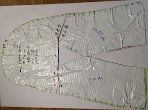 A shape cut out of a duct-tape cast, flattened and stuck to card. Various black marks shoe stitch positions.