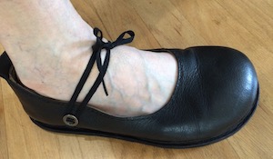 A black leather court shoe with a black shoelace extending from a button below the ankle bone across the foot to the other side of the shoe