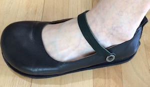 A black leather court shoe with a black leather strap extending from a button below the ankle bone across the foot to the other side of the shoe