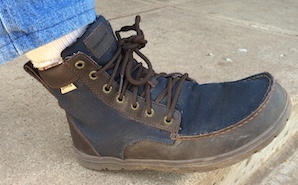 A blue canvas boot with brown leather trim and brown laces, on a right foot clad in cream sock and blue jean leg