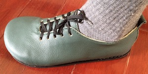 A side-on view of a green leather, flat-soled shoe with black nylon laces and a beige leather lining just visible