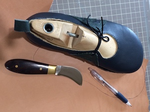 A blue shoe on a wooden last, above a sheet of thick veg-tan leather with a line drawn on it in pen. Also visible a pen and a leather knife