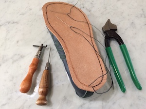 Seen from the bottom, line of stitching sitting in a groove around the perimeter of a veg-tan outsole. Also visible are two needles, a pair of pliers, a groover and a straight awl.