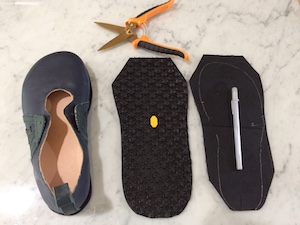 A blue shoe next to two flat pieces of black rubber soling material cut into a rough foot shape. One is lying sole upwards so a Vibram mark is visible. The other is lying sole downwards and has a silver line traced onto it with pen, showing the shape of the leather sole that will be glued to it