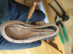 The bottom of a shoe last with a leather welt being attached. A long awl is piercing the welt and the midsole as well as the upper (barely visible).