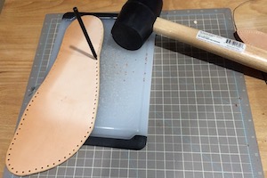 A foot-shaped piece of leather, with a punch and rubber hammer being used to make evenly-spaced hoes around the perimeter, a few mm in from the edge.