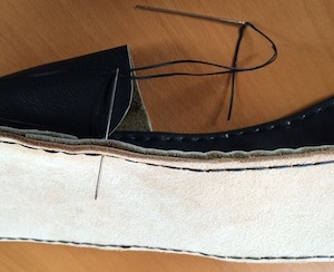 A needle piercing several layers of leather. In the background lies another needle, attached to the same piece of black thread as the first.