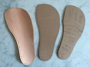 A foot-shaped piece of card with pencil markings on it, a similar shaped piece with holes punched around the edge, and a piece of leather in the same shape.