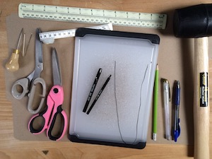 A variety of tools. Rulers, scissors, awl, punches, pencils, pens, needles, thread, rubber mallet, nylon chopping board.