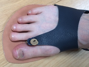 Close-up of a pale-skinned foot in a minimal leather chappal sandal with a 2mm-thick veg-tanned sole and an upper made from dark brown buffalo hide. The sole is attached to the upper between the big toe and the second toe, and at the sides of the foot just forward of the ankles.