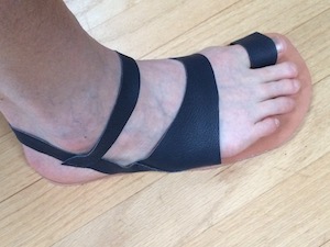 A foot in a sandal that has a loop over the big toe, and straps at the ankle that are attached to the leather sole just forward of the ankle bone. The heel of the foot has slipped sideways off the sole.