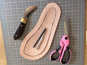 A sole-shaped piece of veg tan leather, surrounded by another of the same shape, side by side with a wood-handled curved knife and a pair of pink-handled fabric scissors.