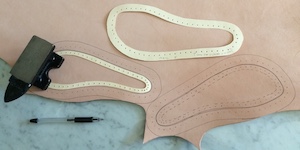 Foot-shaped card patterns on pale veg-tan leather. One of the patterns is being held down with a small iron anvil