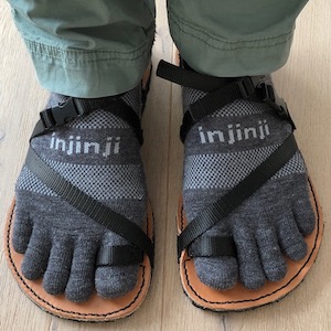 a pair of feet with spread toes, wearing toe socks, seen from above, wearing minimal leather sandals with pale veg-tan leather soles and black nylon straps.