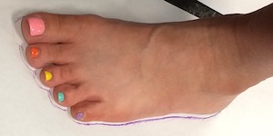 A pale-skinned left foot standing on a white piece of paper with a purple line traced around the edge of the foot. The toenails are painted various colors: pink, red, yellow, green, and purple.