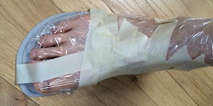 A foot encased in transparent cling wrap standing on a cardboard sole, with pieces of 1 inch masking tape partly wrapped around it