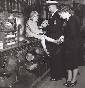 Black and white photo of a man and woman standing at a store counter on which a girl of about 8 years old is sitting. The man is wearing a naval uniform; the woman and girl are in skirts. All are wearing shoes with raised heels.